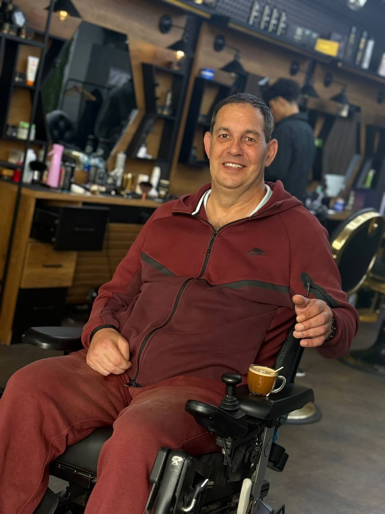 Een klant in een rolstoel lacht breed, met een kop koffie op de armleuning, in de zaak.